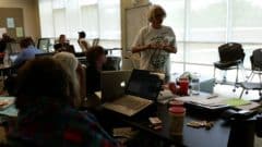 Instructor leads a workshop with participants using laptops seated around a table in a bright classroom.