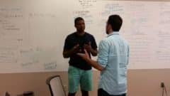 Two men discuss ideas while standing in front of a whiteboard covered with notes and diagrams during a collaborative work session.