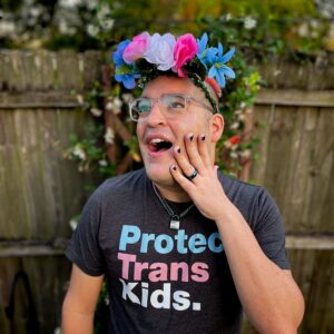 david in a trans floral crown, with painted nails and a trans collar on and a shirt that reads "Protect Trans Kids."