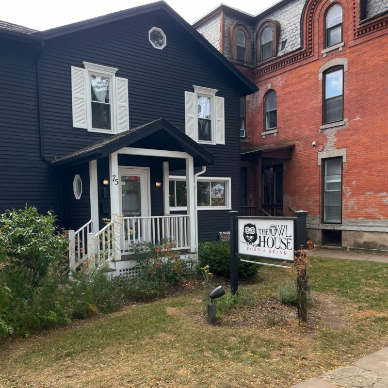 A blue house with a white staircase, porch, and door. The house is a business and has a sign in front of it that says The Owl House Food & Drink.