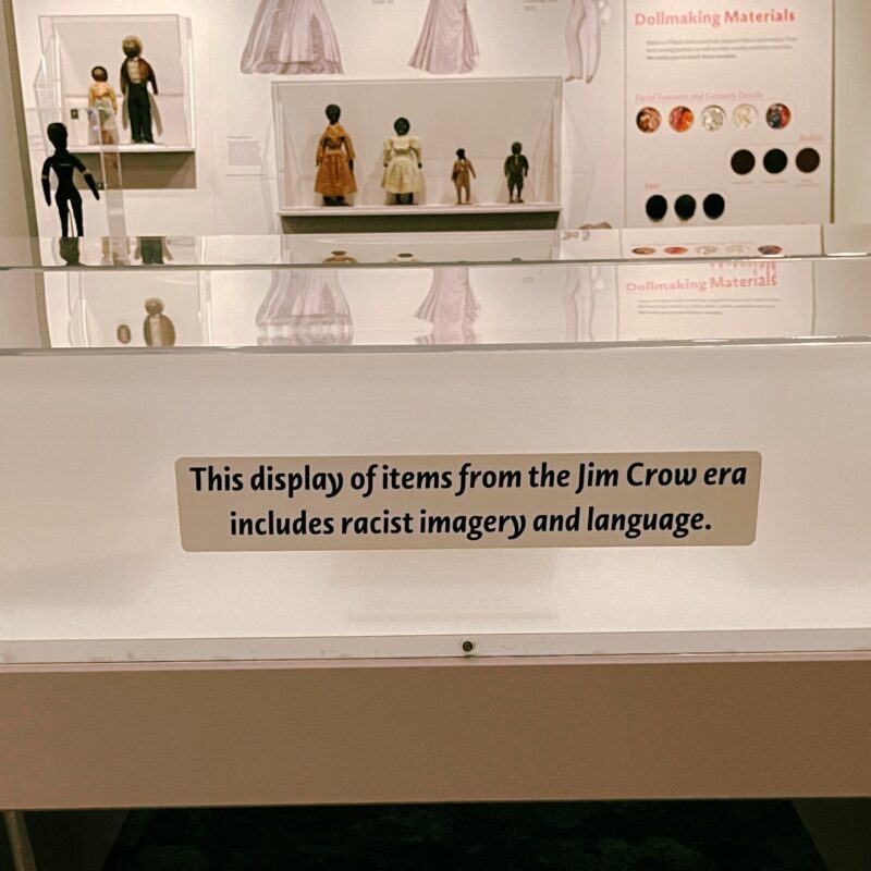 A glass display case that reads, "This display of items from the Jim Crow era includes racist imagery and language." in a room with a variety of black dolls and doll making displays