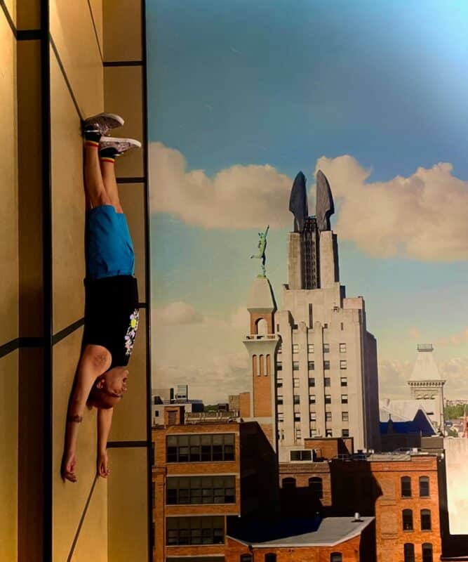 david lying down on the floor in a display that is meant to look like the side of a building, with part of the Rochester skyline in the background