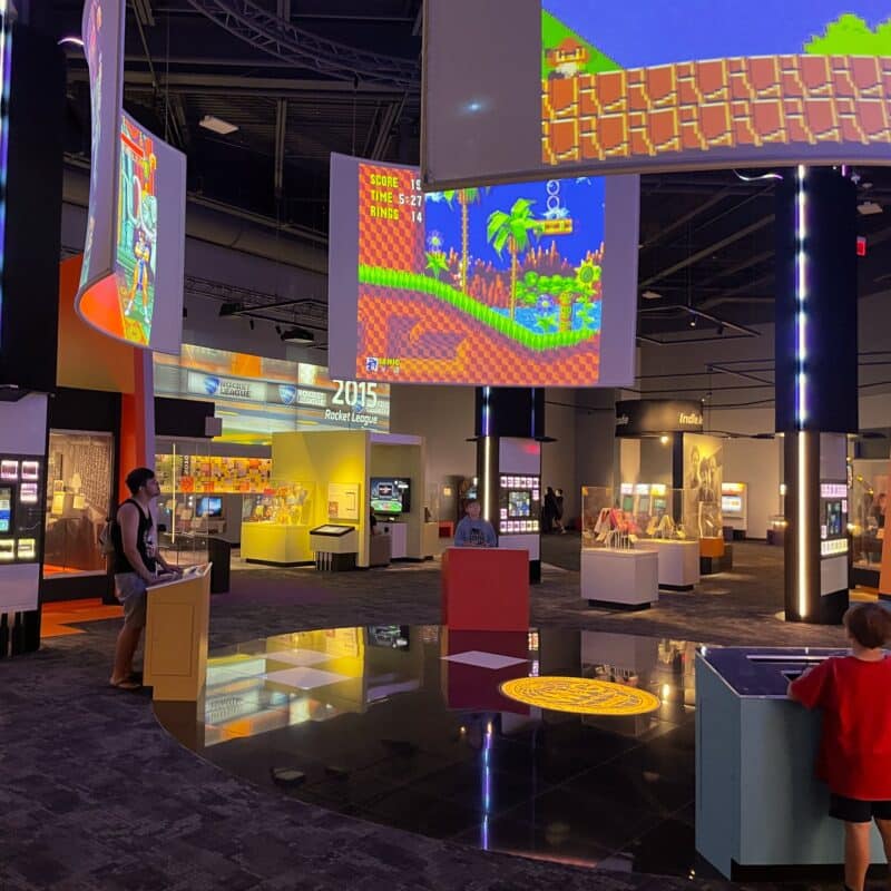 A room of interactive video games at the Strong Museum of Play with people playing games at pedestals on large projector screens