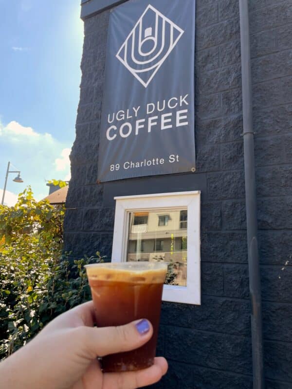 The sign for Ugly Duck Coffee in Rochester, NY, with a hand with purple painted nails holding a ginger beer espresso drink in the foreground