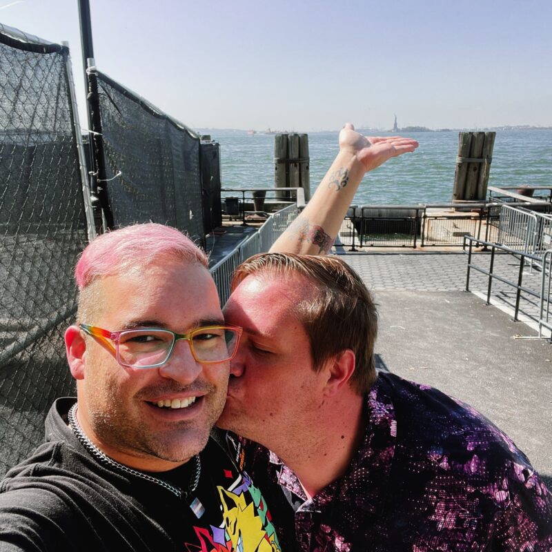 A selfie of david and their husband kissing their cheek. david is holding their palm up to look like they are holding the Statue of Liberty in the background