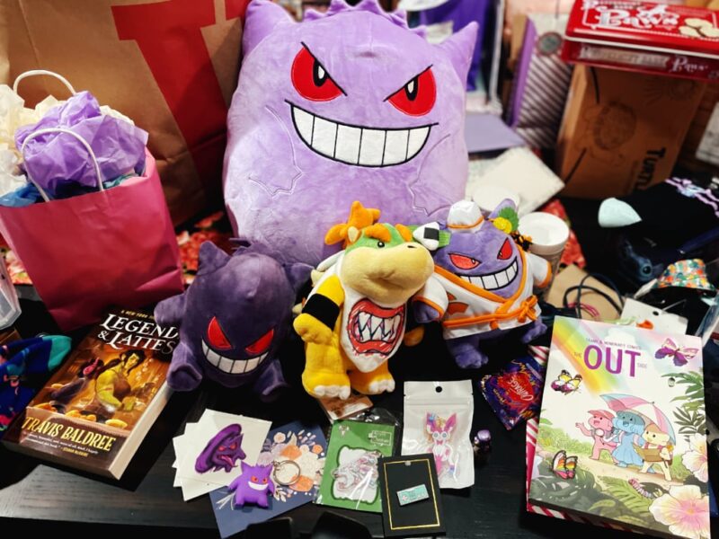 A collection of holiday gifts including Gengar and Bowser Jr plushes, the book Legends & Lattes, the comic anthology The Out Side, and Sylveon and Gengar trinkets