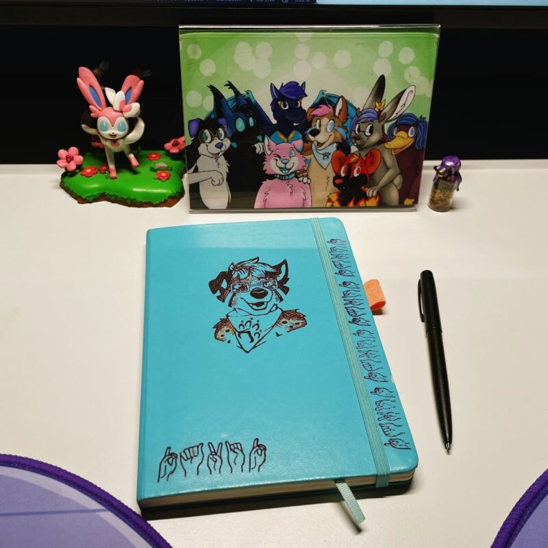 A blue notebook next to a pen and in front of a framed furry family portrait, Sylveon statue, and bottle of herbs. The notebook has an engraving of the fursona Bowie on it, and several versions of the name david in ASL