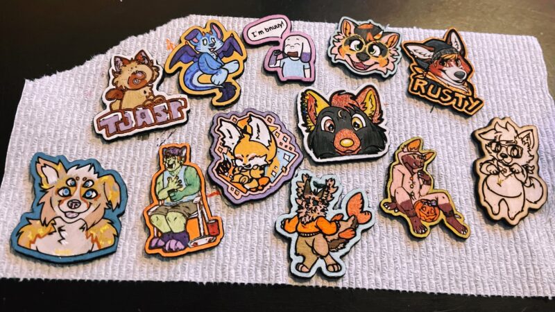 A variety of fursona art wooden pins that are painted but not varnished on a paper towel