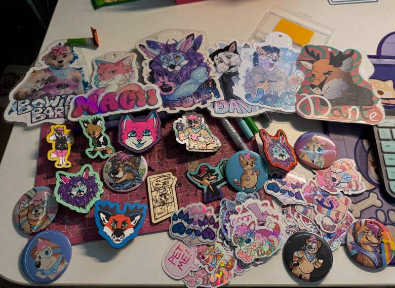 A collection of printed furry badges, painted wooden furry pins, fursona buttons and stickers, and markers on a desk in a cluttered, scattered fashion.