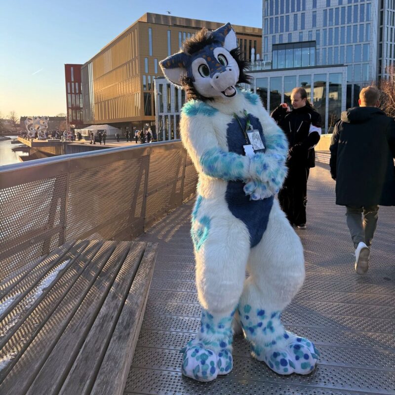 A person in a fluffy, blue and white anthropomorphic animal costume stands on a bridge, posing playfully. The fur suit features large ears, a friendly facial expression, and spotted patterns. The background shows modern multi-story buildings under a clear blue sky. Other people walk nearby, adding to a lively urban atmosphere. Sunlight casts shadows across the bridge
