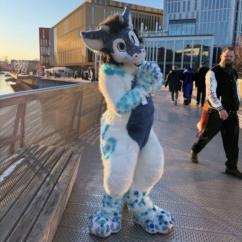 A person in a blue and white furry costume resembling an anthropomorphic creature stands on a modern metal walkway. The costume features large ears, a patterned face, and fluffy paws. The background shows a waterfront area with modern architecture, including glass-fronted and colorful buildings. Several people in various costumes and casual attire walk in the background, enjoying the sunny day