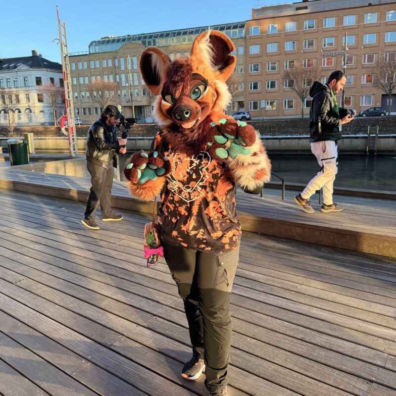 A person is dressed in a brown and orange furry costume with large ears and green paw pads, standing on a wooden deck. The outfit features a stylized face on the shirt with orange and black patterns. In the background, two other people are visible. One is holding a camera, and the other is looking at their phone. They are near a