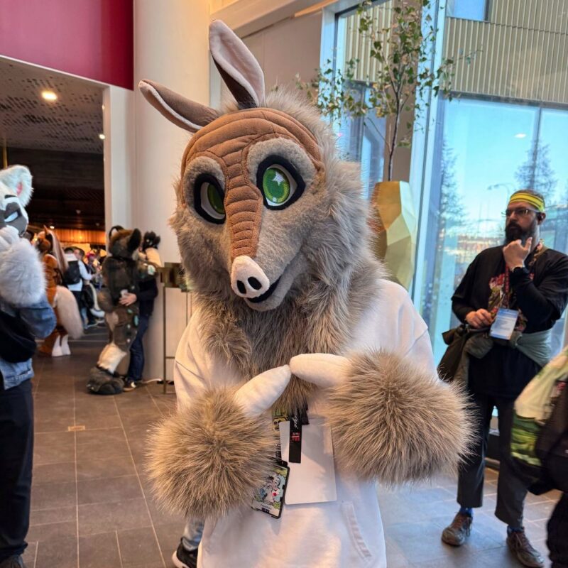 A person wearing a furry costume with large ears, a long snout, and vibrant green eyes stands indoors. The costume is predominantly gray and white, with fluffy fur and a large head resembling an aardvark. They are holding a badge or card with a lanyard. Behind them, several people, some also in animal costumes, are ming