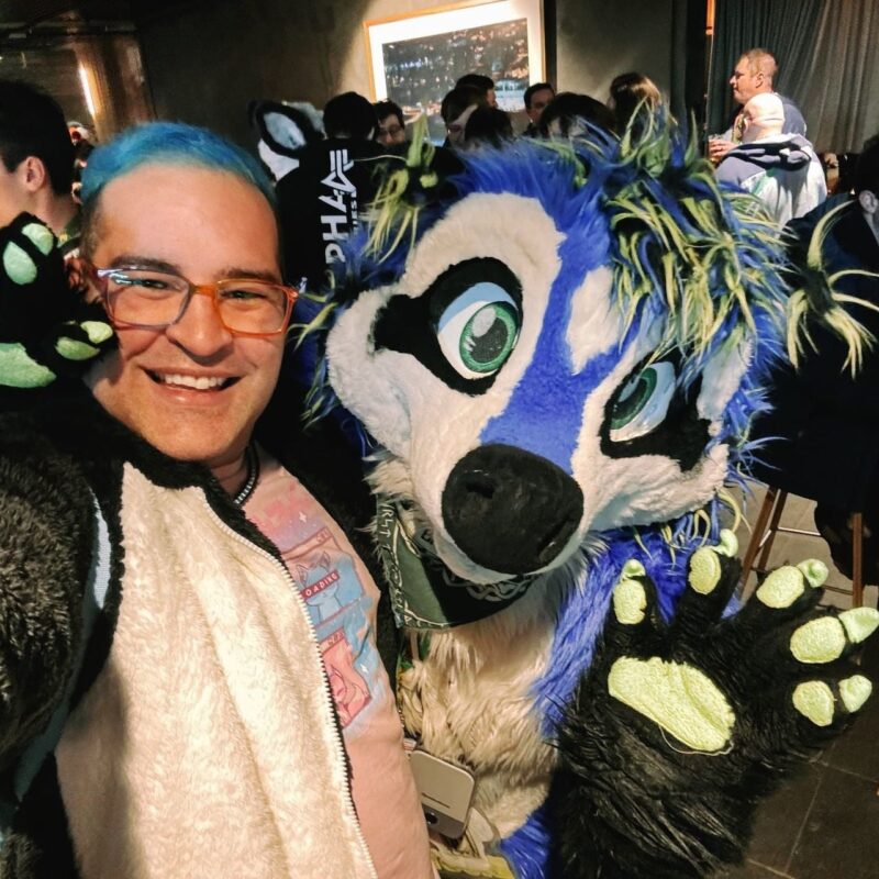 A person with blue hair and glasses is smiling alongside someone in an elaborate furry costume. The costume is predominantly blue and white, featuring bright green eyes, a black nose, and neon green paw pads. The setting appears to be a lively indoor gathering or event, with several people visible in the background, some engaging in conversations. The atmosphere seems festive and