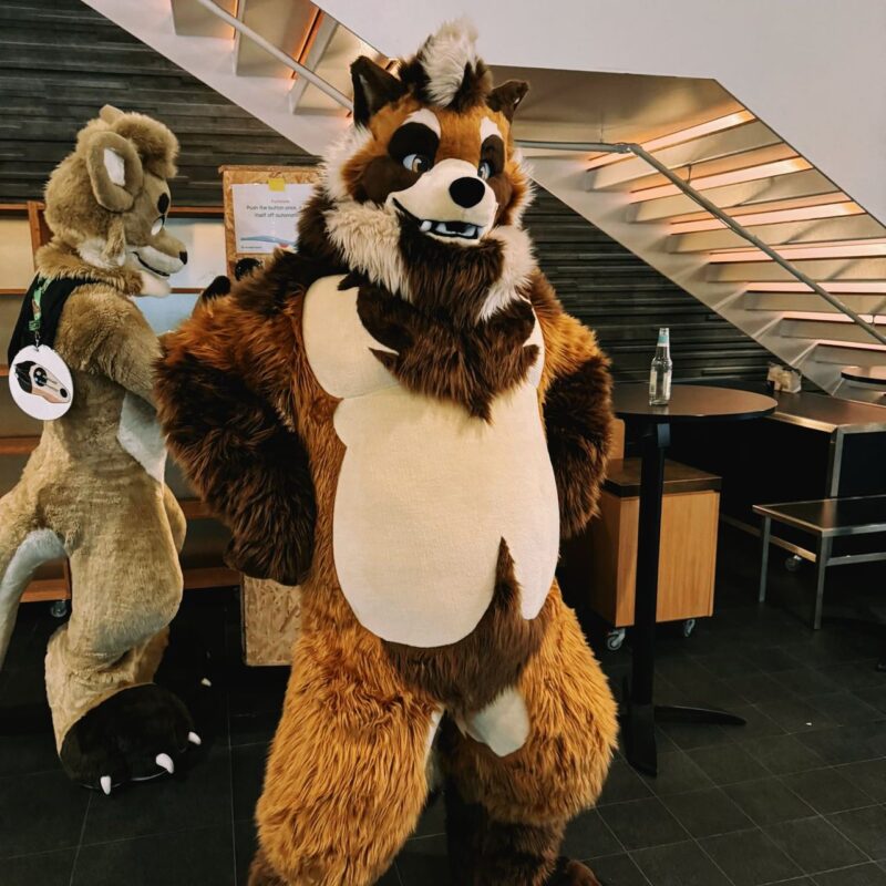 A person in a detailed furry costume stands indoors near a staircase. The costume is brown and white, resembling a stylized animal with a large head, fluffy mane, and prominent eyes, exuding a playful expression. Another person in a lighter brown furry outfit is in the background, partially turned away. A round table with a glass bottle is nearby,