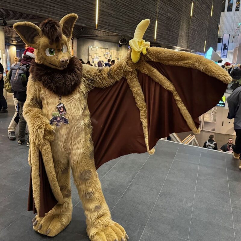 A person in a furry costume resembling a bat is standing indoors. The costume is brown with large ears, green eyes, and a big furry mane. The outfit features extended wings, with one hand holding a peeled banana. The venue has a modern design, with people visible in the background, some wearing backpacks. The scene appears lively, suggesting a convention