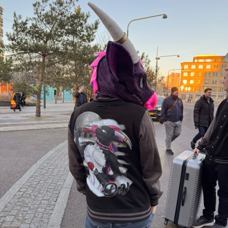 A person is walking outdoors wearing a purple creature mask with pink accents and horn details. They are dressed in a dark jacket featuring a colorful fantasy creature design on the back, holding a rose. The background shows a city street with a few trees, people walking, and buildings reflecting the sunset. A person nearby is pulling a large silver suitcase. The overall