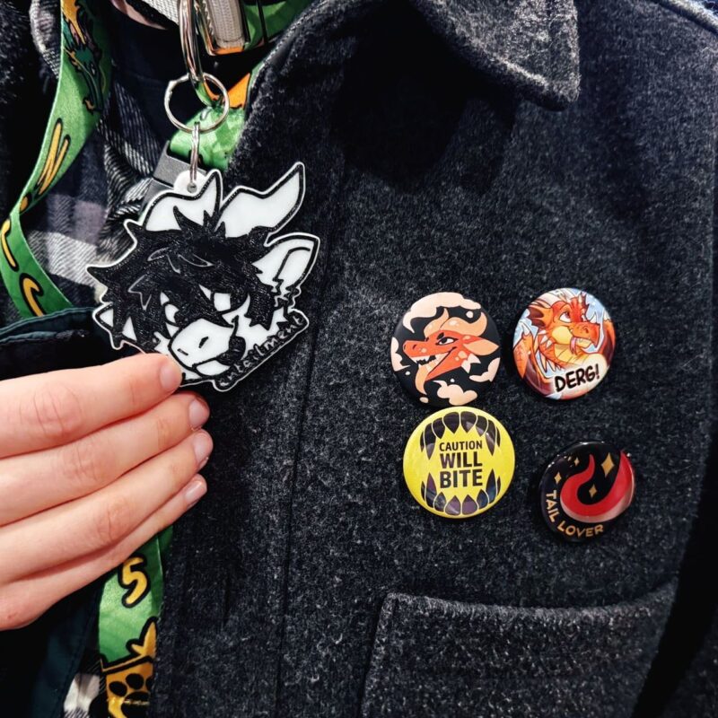 A person wearing a dark gray coat is displaying several colorful buttons on the chest. The buttons feature cartoonish dragon designs with captions like DERG!, CAUTION WILL BITE, and TAIL LOVER. Nearby, a hand is holding a keychain with a black and white illustration of a dragon head. A green lanyard with yellow