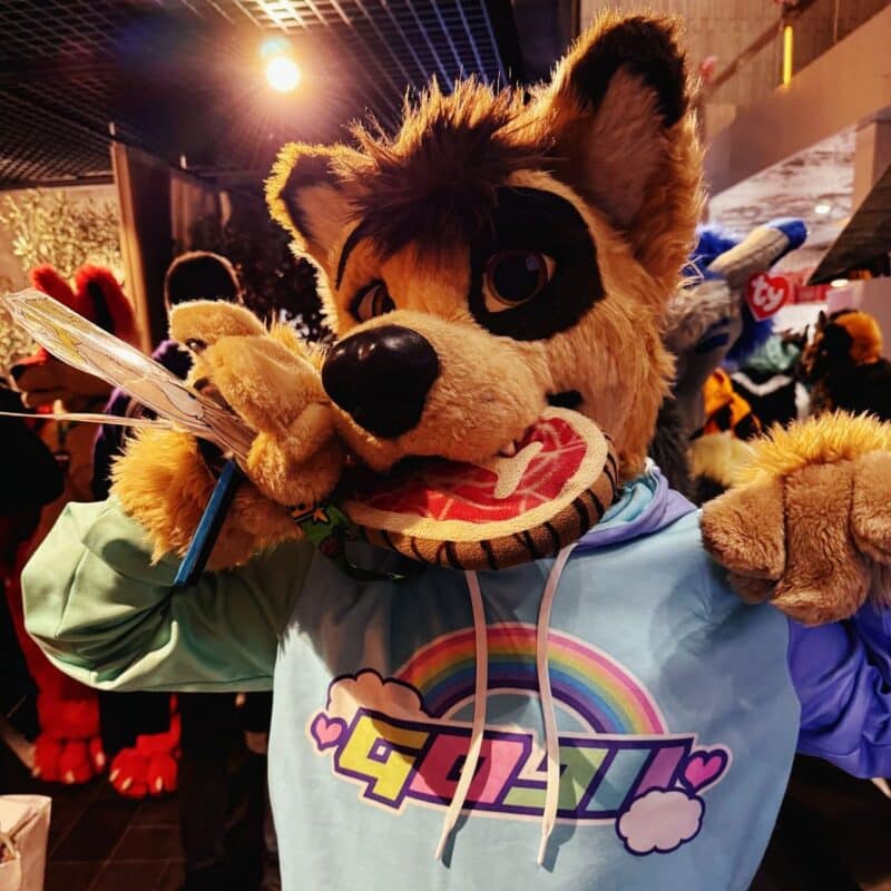 A person in a furry costume is posing indoors. The costume resembles a cartoonish dog with tan fur, large eyes, and a bushy tail. They are holding a round, red-and-white fan in their mouth. The costume includes a colorful hoodie featuring a rainbow design and stylized text. Other people in costumes are visible in the background, along