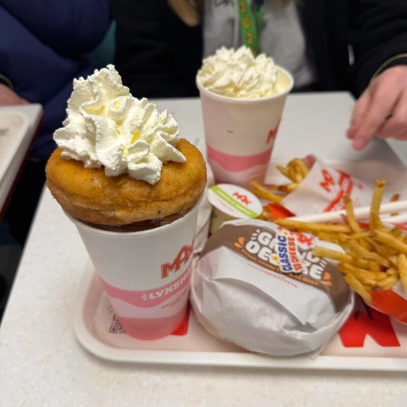 A tray with fast food items, including a burger wrapped in branded paper, a pile of French fries, and two pink-striped cups topped with whipped cream. One of the cups has a donut placed on top, also topped with whipped cream. The tray is set on a table, and a person with long hair wearing a black jacket is seated nearby,