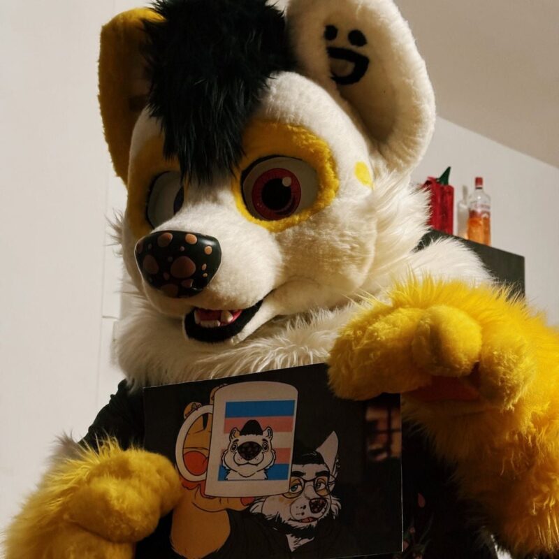 A person in a large, fluffy fursuit is showing a poster. The fursuit has a yellow and white color scheme with a playful expression. The ears have a smiley face detail inside. The poster depicts cartoon characters, including a cup with a stylized animal face featuring the colors of the transgender pride flag in the background. The environment