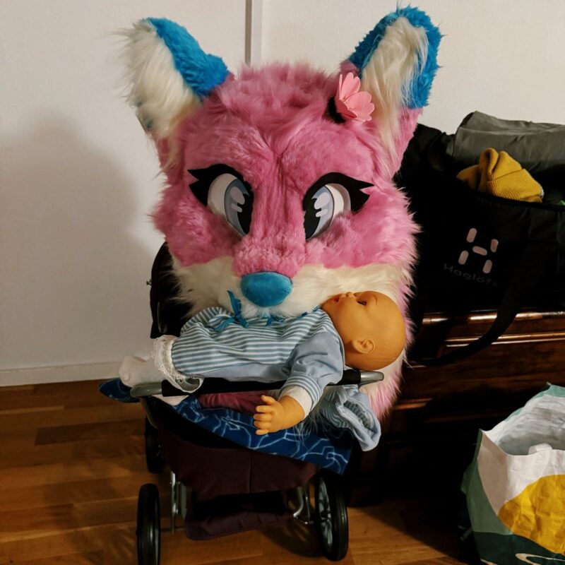 A large, colorful pink and blue furry head resembling a cartoonish animal is resting on a baby stroller. The plush head has large eyes and a flower accessory. In the stroller, there is a doll dressed in a stripped blue outfit. Behind the stroller, there are bags on the floor and against a wall, including one that is black with the word
