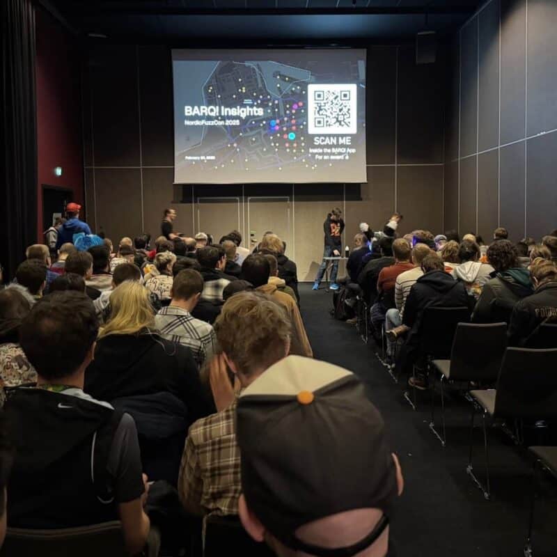 A large group of people is seated in a conference room facing a presentation screen. The screen displays the title BARQ! Insights - NordicFuzzCon 2025 with a QR code beside it. The audience appears engaged, with some wearing casual clothing and a few with colorful hairstyles. The room is dimly lit, and two people are