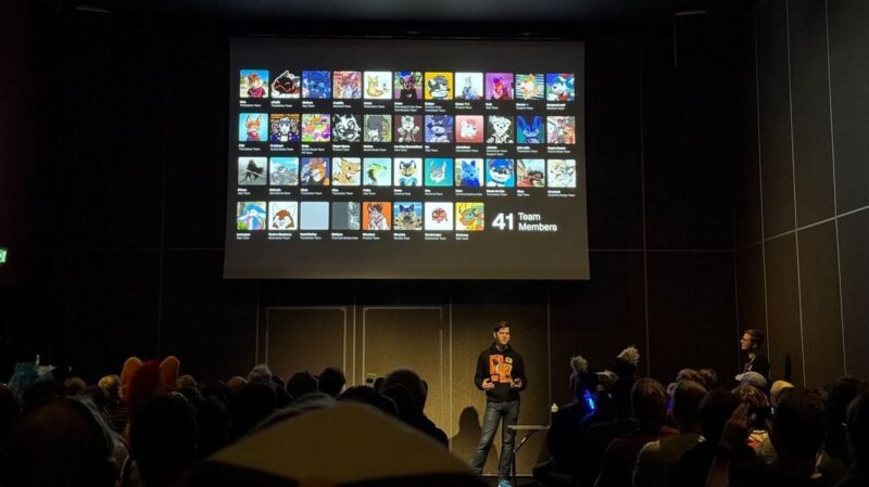 A person stands on a stage in a dimly lit room, presenting to an audience. They are in front of a large screen that displays cartoon-style avatars in a grid, representing team members. The text on the screen reads “41 Team Members.” The audience is seated, with several individuals wearing costumes or headbands featuring animal ears. The atmosphere appears