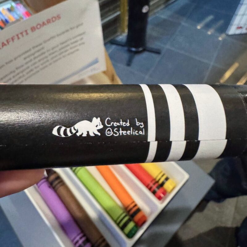 Close-up of a black object with white stripes, featuring a small silhouette of a raccoon next to the words Created by @Steelical. In the background, there is a display with colorful cylindrical objects. A sign titled GRAFFITT BOARDS is partially visible. The setting appears to be indoors, with a tiled floor and