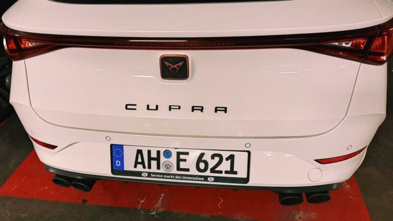 The image shows the rear view of a white Cupra car, parked indoors. The car features sleek tail lights and a distinct brand emblem above the word CUPRA on the trunk. The license plate reads AH IE 621, with European and registration symbols. The car is positioned on a surface with red markings. Dual exhaust pipes are