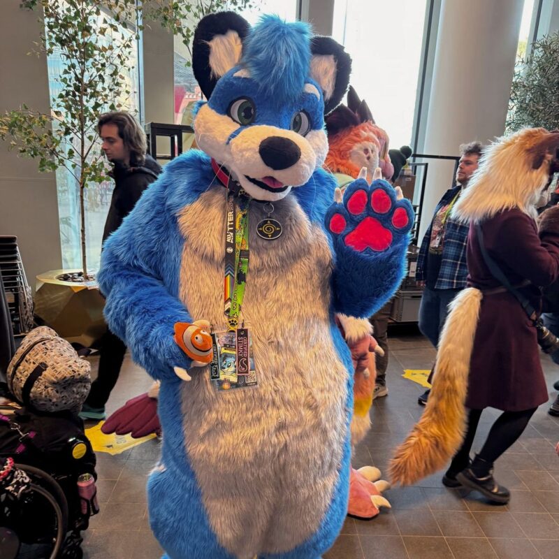 A person dressed in a bright blue and white furry costume with black ears and a pink paw pad on their raised hand. They wear a lanyard with badges around their neck and hold a small stuffed clownfish in one hand. The setting appears to be a convention or gathering with other attendees wearing costumes and casual clothing in the background. The environment is