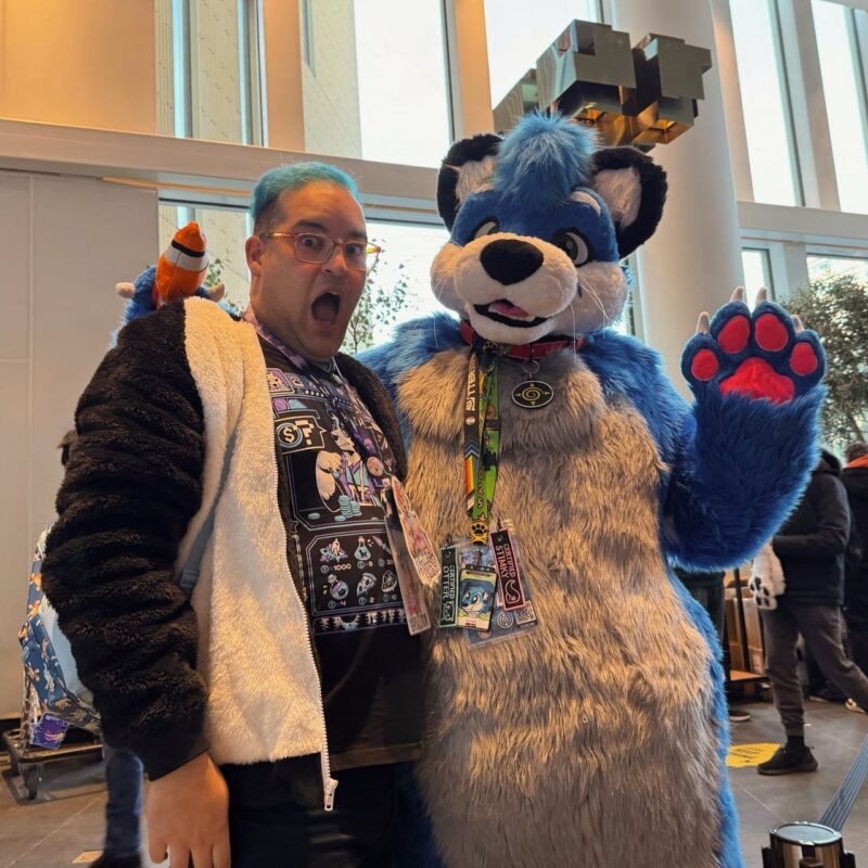 A person with blue hair, wearing glasses and a fluffy black and white jacket, stands excitedly beside someone in a blue and grey animal costume with black ears and a large pink paw. Both appear to be enjoying a convention or event inside a bright, glass-walled building. The costumed individual wears various colorful badges around their neck, and the person