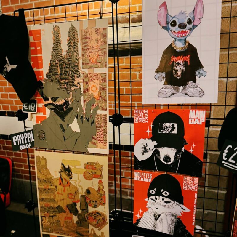 A display of urban and cartoon-style art pieces on a black grid frame. The left side features two intricate, abstract cityscape posters. The right side includes a blue cartoon character in a black band shirt, and below it, a black and white drawing of a person in a cap labeled MAW CAP on an orange background. Below, a