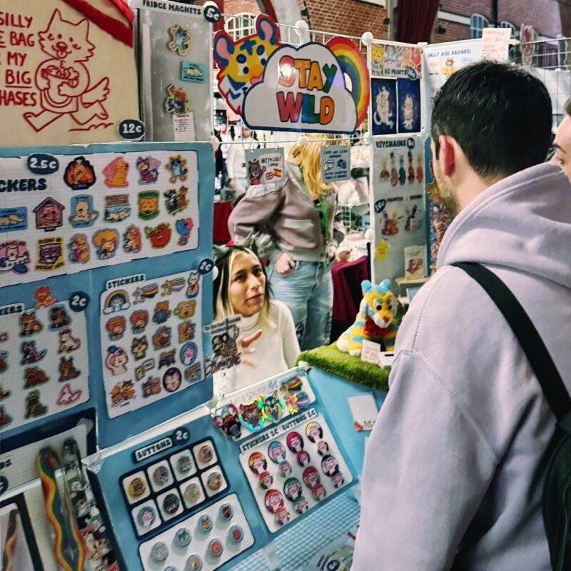 A market stall displays an array of colorful stickers, badges, and magnets with cartoon characters and designs. A vendor, standing behind the counter, converses with a customer dressed in a light hoodie looking at the items. A plush toy tiger sits on artificial grass as decoration. A rainbow sign with the words STAY WILD hangs above. The