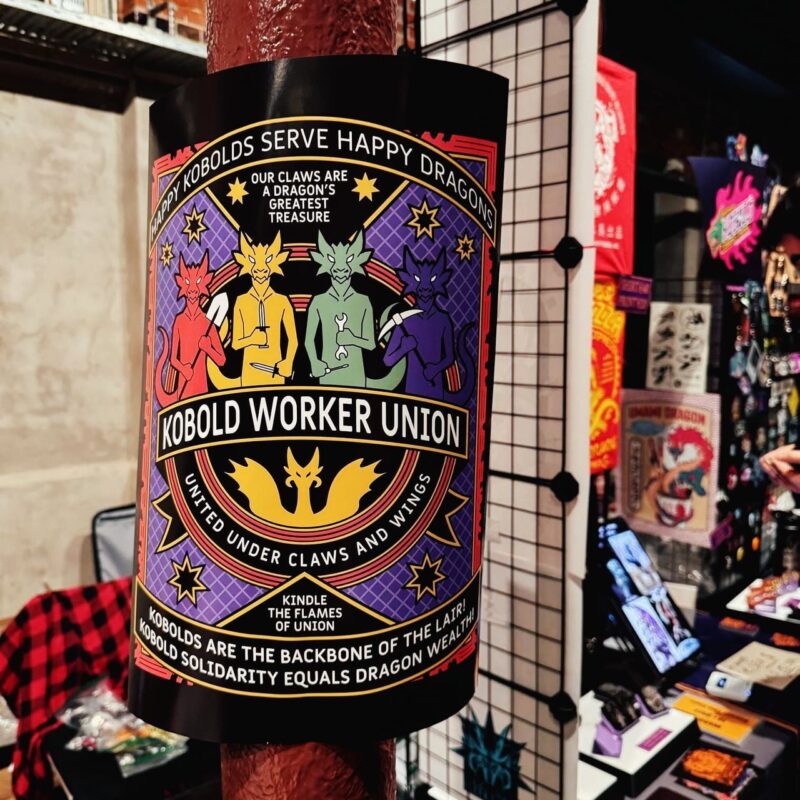 A poster titled Kobold Worker Union is taped to a pole. It features colorful illustrations of five kobolds, each a different color, holding tools. The text includes slogans like Happy Kobolds Serve Happy Dragons, Our Claws are a Dragon's Greatest Treasure, and United Under Claws and Wings. The bottom section reads