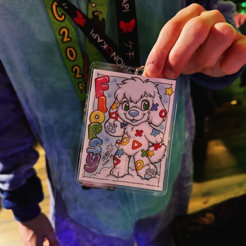A person is holding up a colorful badge featuring a cartoon character with white fur, large green eyes, and a playful smile. The character is covered in multicolored, playful X and O symbols. The word Flopsy is written vertically in bold, colorful letters to the left. The person appears to be wearing a blue outfit, and a