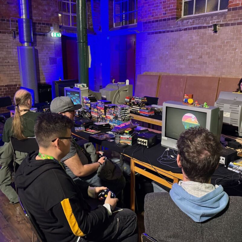 A group of people are seated in front of retro video game setups in a room with exposed brick walls and blue lighting. Various old televisions display colorful game graphics. Stacks of video game cartridges and consoles are arranged on tables. One person is holding a controller, while others appear to be engaged in gameplay. The atmosphere is nostalgic, showcasing classic gaming culture