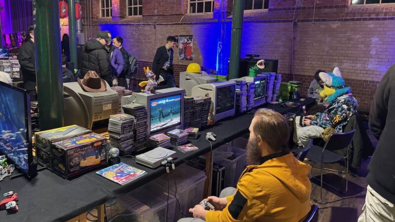 The image depicts a retro gaming event in a dimly lit room with exposed brick walls. Multiple CRT monitors display video games, with stacks of game discs beside them. A person in a yellow jacket is playing on a classic console. Another person in a colorful furry costume is sitting nearby, also engaged in gaming. Several people are standing and walking around the
