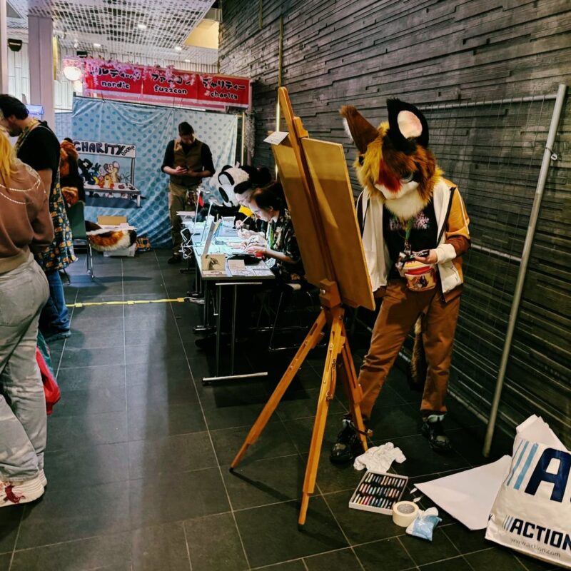 A person dressed in a fox costume with large ears and a colorful face is painting at an easel in a convention or event setting. They are surrounded by art supplies, including pastels and paper on the floor. Nearby, a person is seated at a table with electronic devices. In the background, several people are engaged in activities near a decorated booth