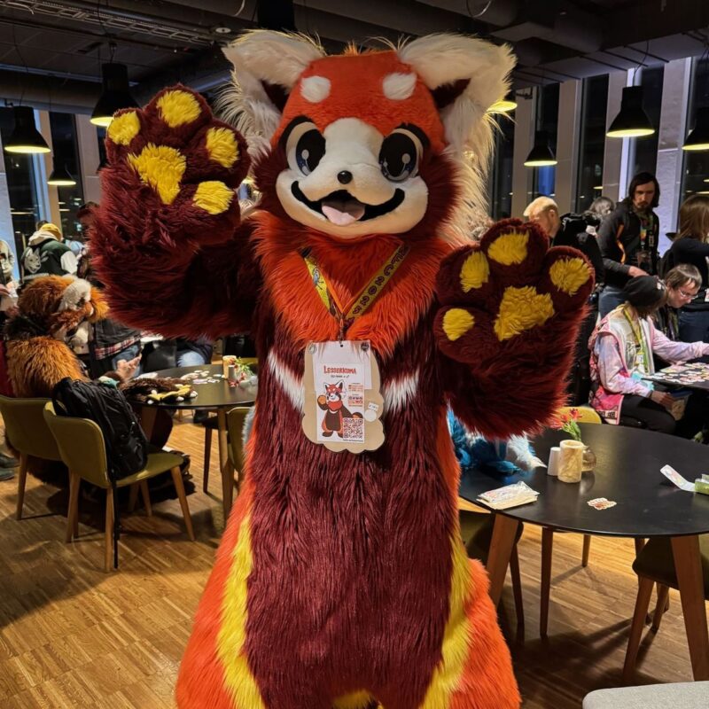 A person wearing a bright, furry costume resembling a red panda is posing with paws upraised in a cheerful manner. The costume features red, white, and orange fur, with large, expressive eyes and a smiling mouth. The individual has a badge around their neck. In the background, people are gathered in a cozy indoor setting with tables and chairs,