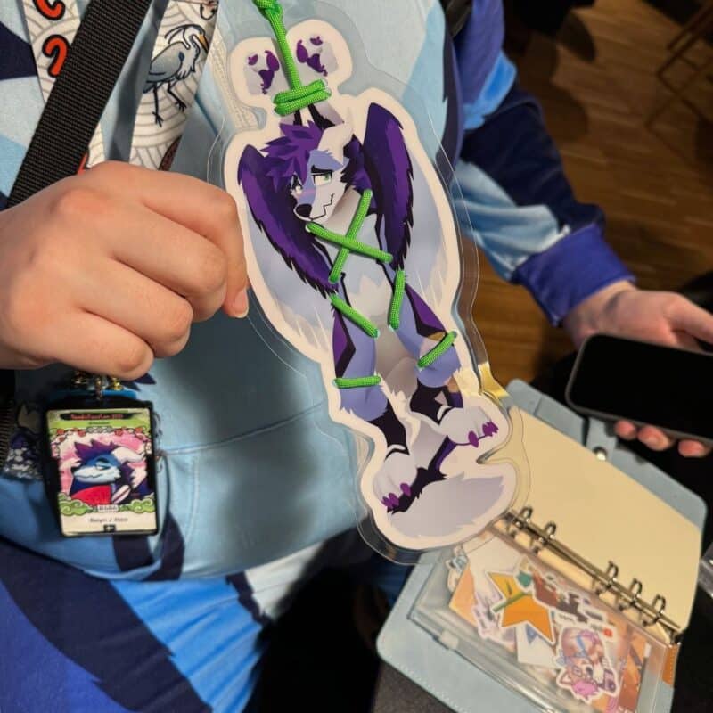 A person wearing a blue hoodie and holding a laminated card with an anime-style character, featuring a purple and white furry creature with green ropes tied around it. They're holding the card in one hand and a smartphone and open binder in the other. The binder contains colorful illustrations and the person is wearing a badge with a similar character image. The scene seems to