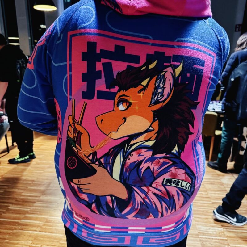 The image shows a person's back wearing a colorful hoodie. The hoodie features a vibrant anime-style illustration of a dragon-like character holding a bowl of ramen with chopsticks. The character is adorned in a patterned robe and has a playful expression. The background of the design is pink, with a mix of geometric shapes and Asian script. The setting appears to be
