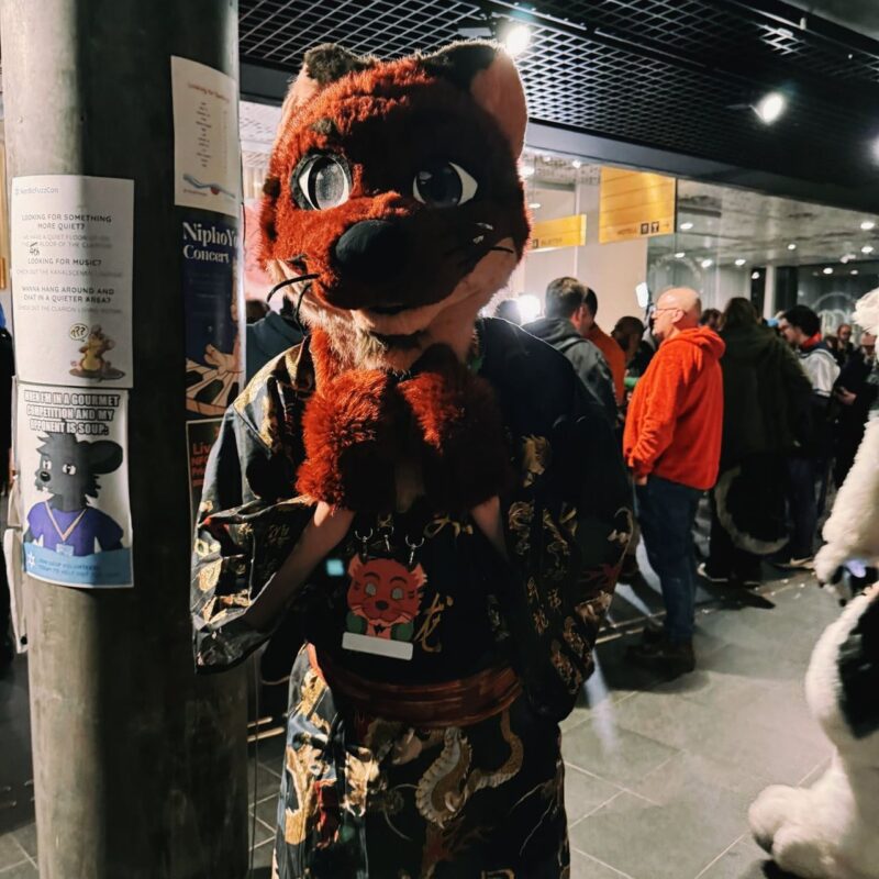 A person dressed in an intricate, red fox costume stands indoors among a crowd. The costume includes a large fox head with expressive eyes and a dark, patterned robe featuring gold details. The individual is posing with their paws together near their chin. Behind them, several people in casual clothing are visible, and there are posters on a nearby pillar. The scene