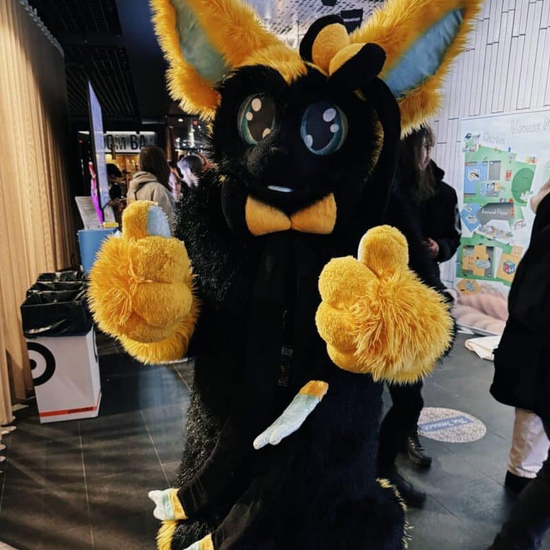 A person in a black and yellow fursuit with large ears and big eyes is giving two thumbs up. They are wearing a matching yellow bow tie. The background shows people and a colorful map on a wall. The scene appears to be indoors, possibly at an event or convention, given the casual, festive atmosphere.