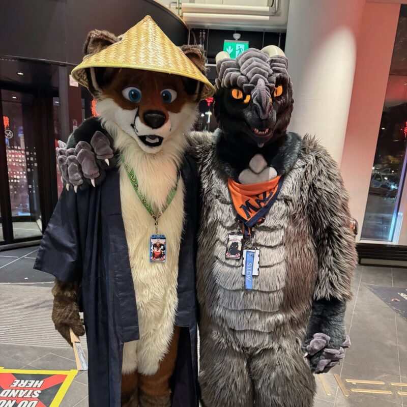 Two people in animal costumes stand together indoors. The left person wears a fox costume with a straw hat, blue robe, and a lanyard with an ID badge. The right person wears a dinosaur-like costume with gray fur, an orange bandana, and a lanyard with a badge. Both are smiling, and their arms are around each