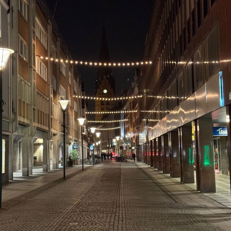 A cobblestone street at night features strings of warm, glowing lights suspended above. The buildings on either side are illuminated by street lamps, creating a cozy atmosphere. In the distance, a tall spire with a clock is visible against the night sky. The street appears quiet with a few pedestrians and bicycles. The scene gives a sense of calm urban