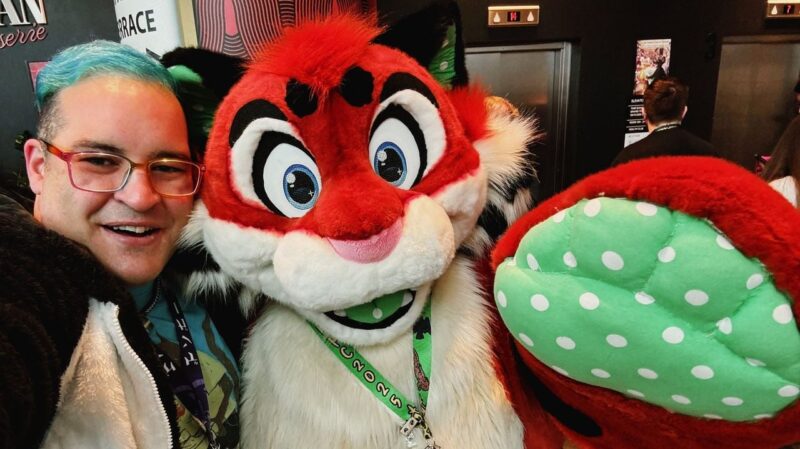 A person with blue hair and glasses smiles next to a large, colorful furry mascot. The mascot features red and white fur, large black eyes with blue highlights, and a wide smile with its mouth slightly open. Its left paw, prominently displayed in the foreground, is green with white dots. The person is wearing a lanyard and a black and