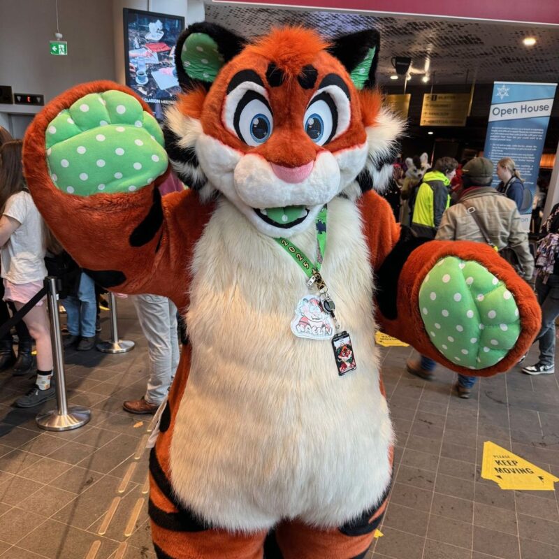 A person dressed in an orange and white tiger costume with large cartoonish eyes is standing in a crowded indoor setting. The costume features green paw pads with white polka dots, and a lanyard with badges hangs around the neck. In the background, there are people milling about, with visible signs and banners, including one that says Open House
