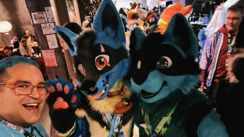 A person with blue hair and glasses is smiling widely next to two people wearing blue and black animal costumes with large ears and expressive eyes. One costume includes a collar with a tag reading Strike. They are at a crowded event with other attendees in various outfits visible in the background. Posters are seen on the wall to the left, and there is a
