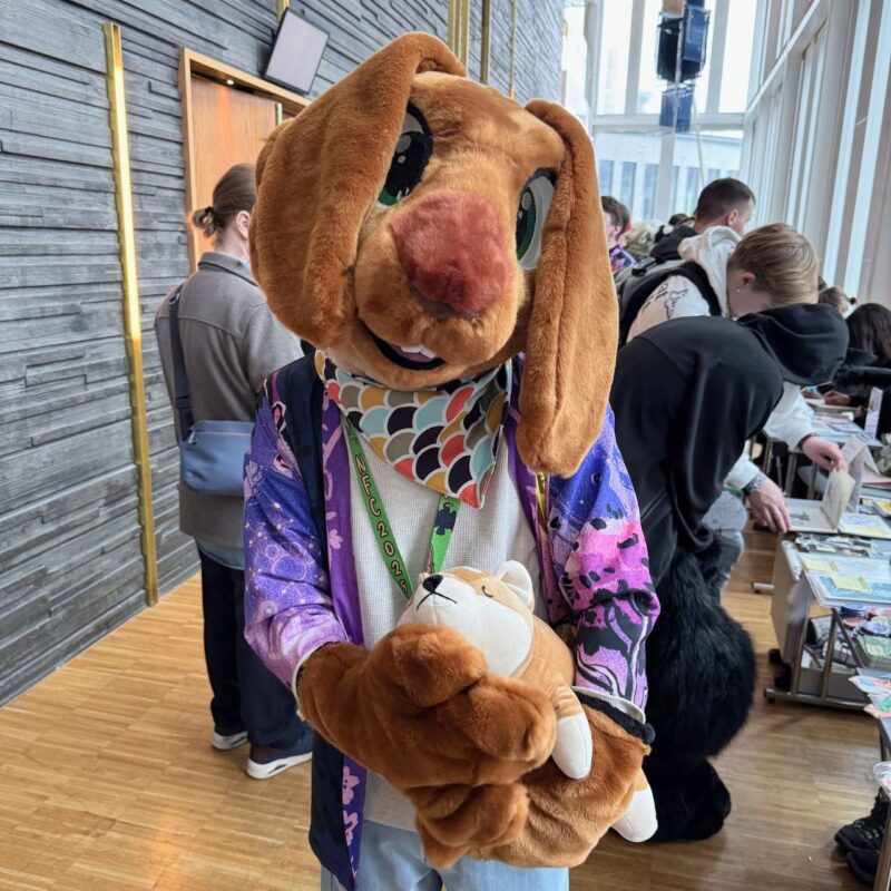 A person is wearing a brown rabbit costume with large green eyes and long ears. They are holding a small, plush toy that resembles a white bear. The costume includes a colorful scarf with patterns and a purple jacket with various designs. The scene appears to be at a convention or event, as there are other people in the background looking at tables with items
